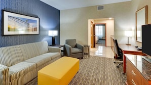 A hotel room with a patterned sofa, a yellow ottoman, a desk with a TV, and a framed picture on the wall. - Drury Inn & Suites Sikeston, MO (Sikeston)