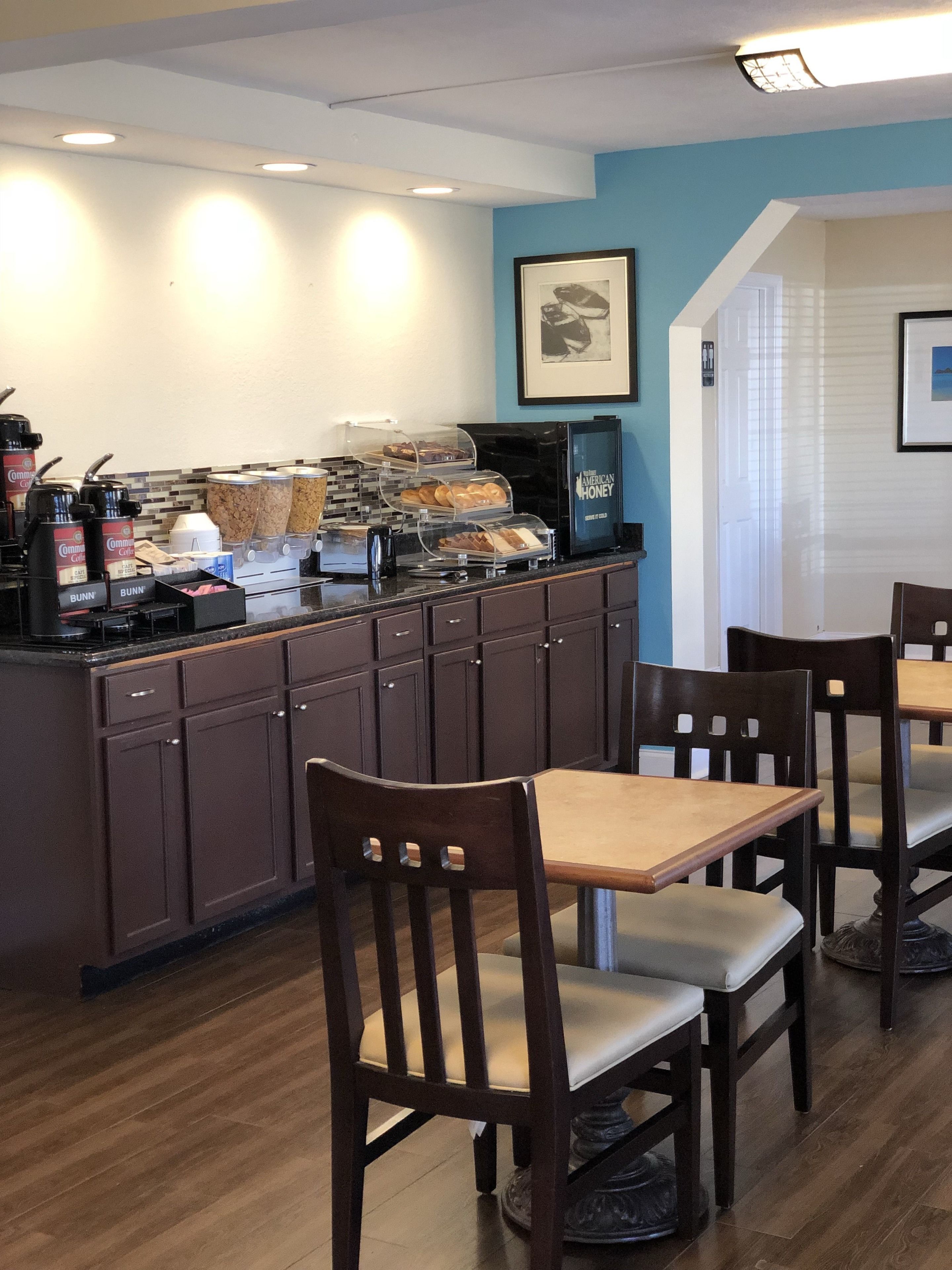 Un café con sillas y mesas de madera, una estación de café con varias máquinas y recipientes, y un cuadro en la pared.