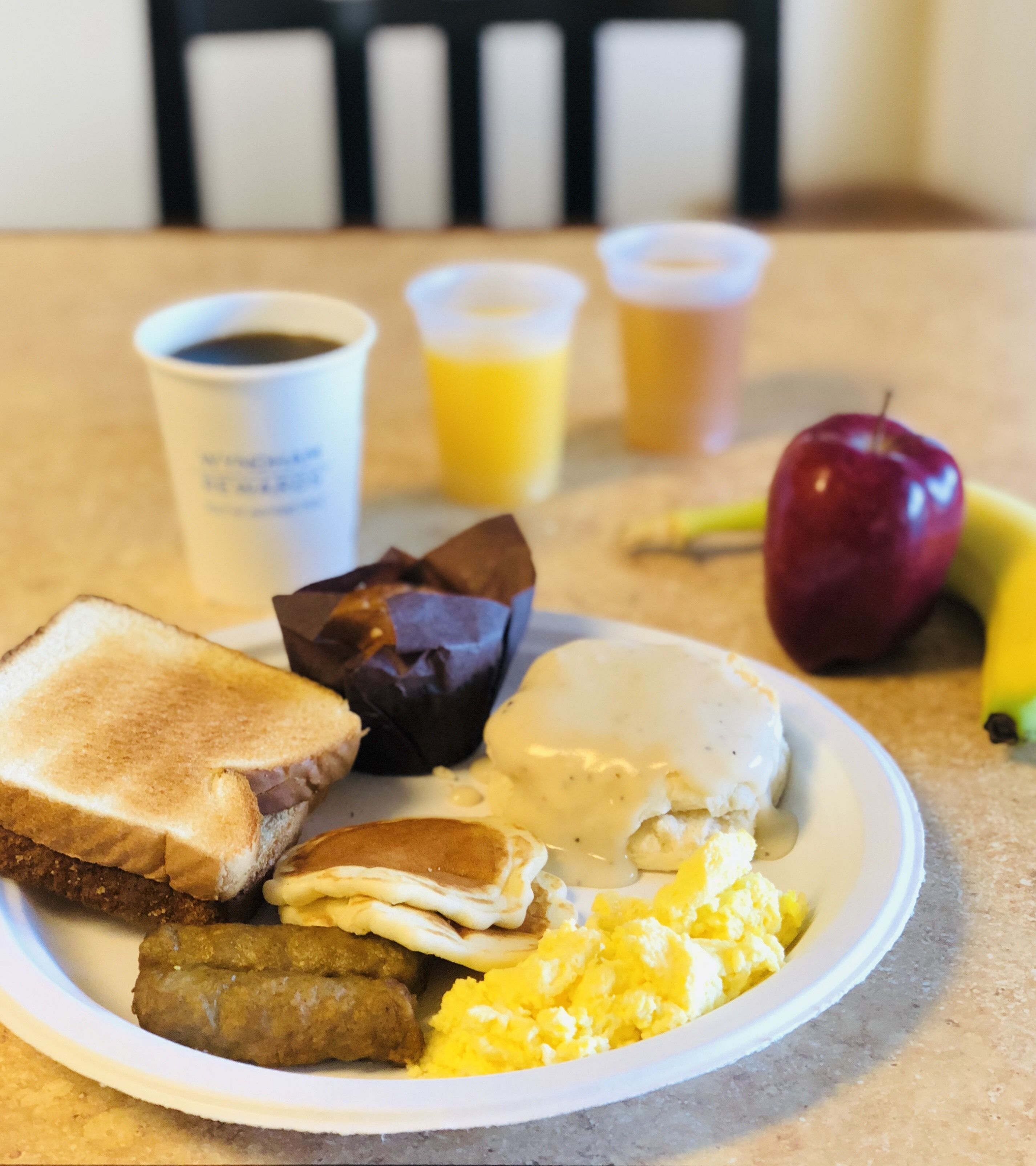 A plate of breakfast food including toast, eggs, sausage, and scrambled eggs, with drinks and an apple on the table.