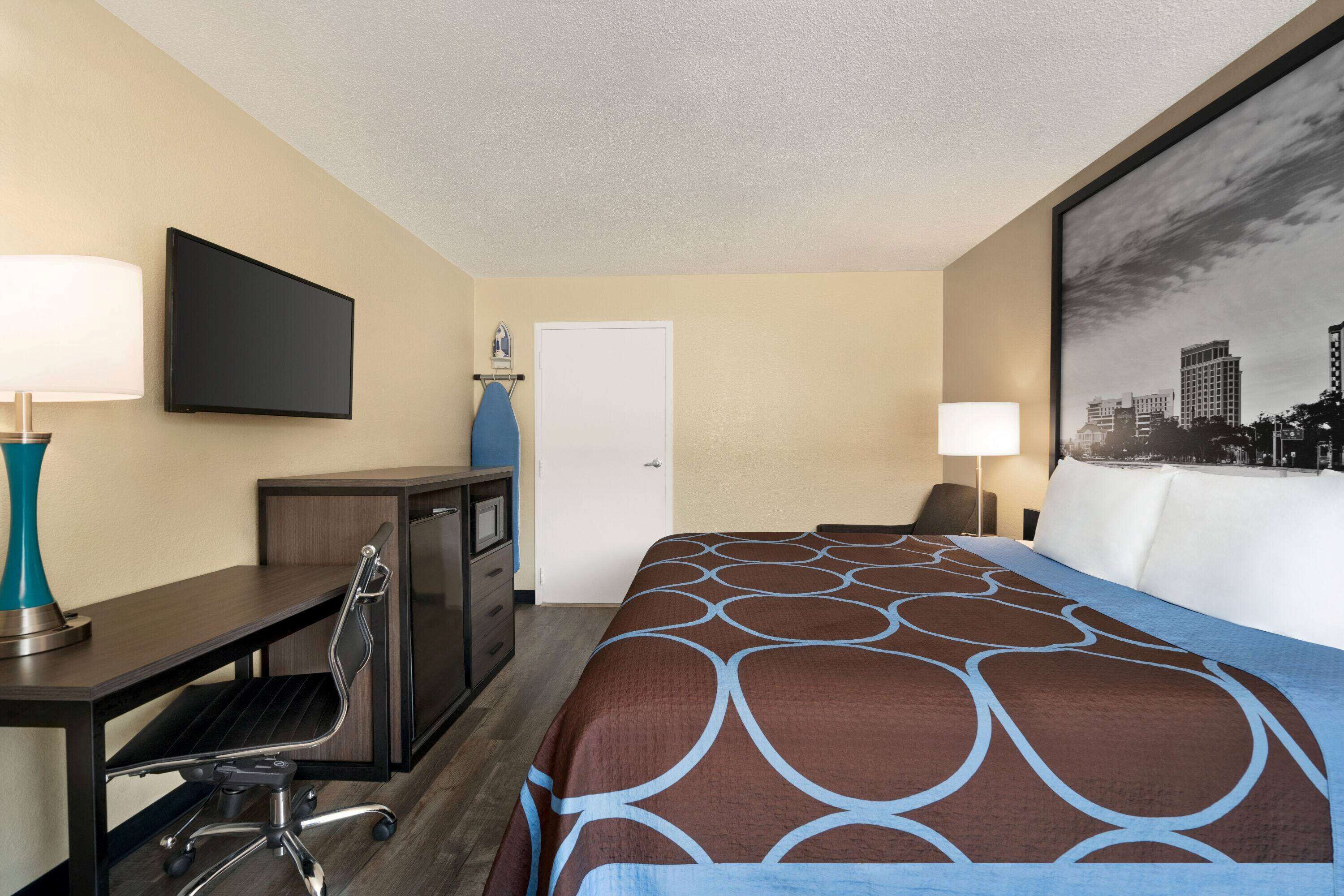A hotel room with a bed, desk, chair, TV, and a painting on the wall.