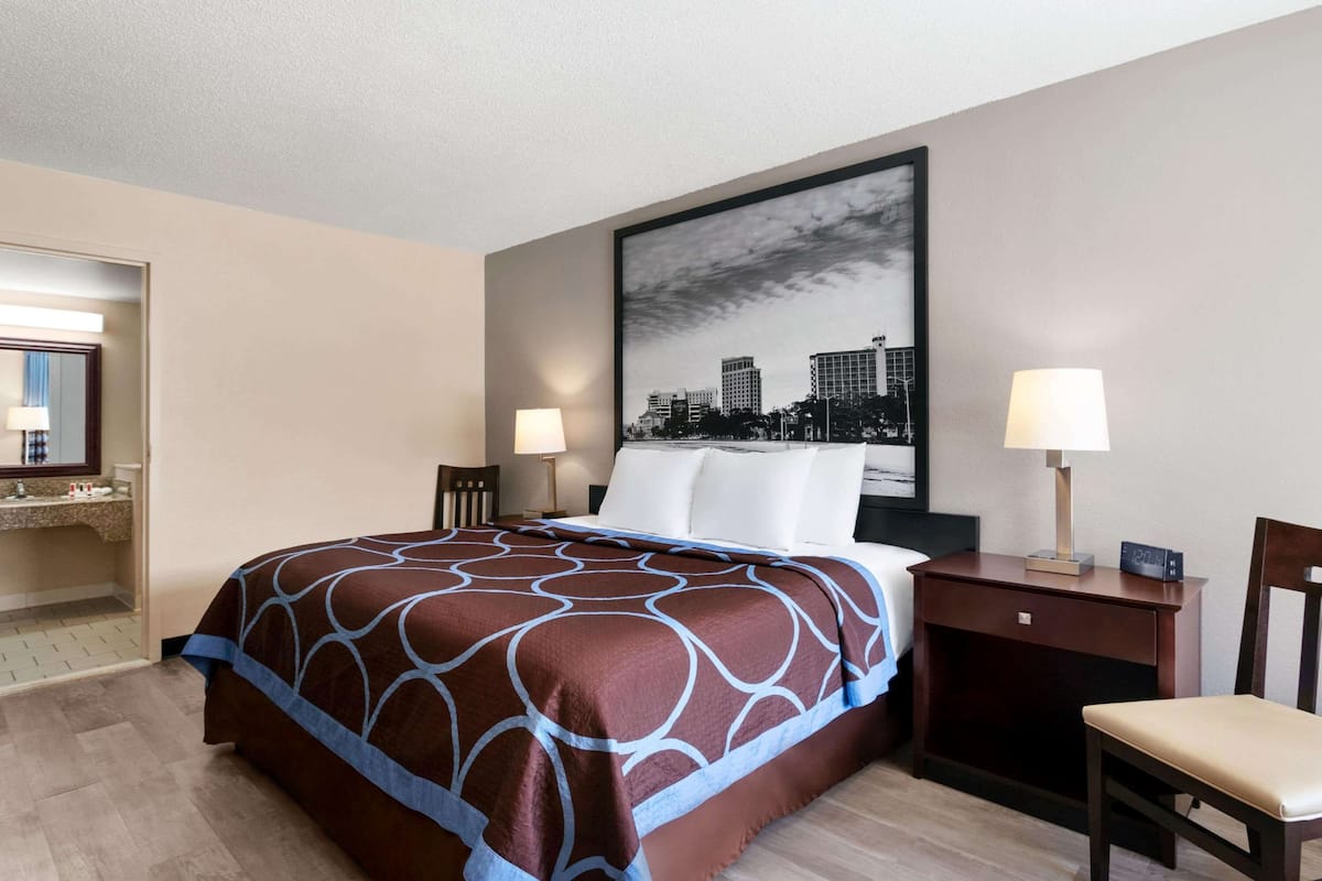 Habitación de hotel con una cama grande, dos mesitas de noche, una silla, un espejo y una obra de arte con vista a la ciudad.