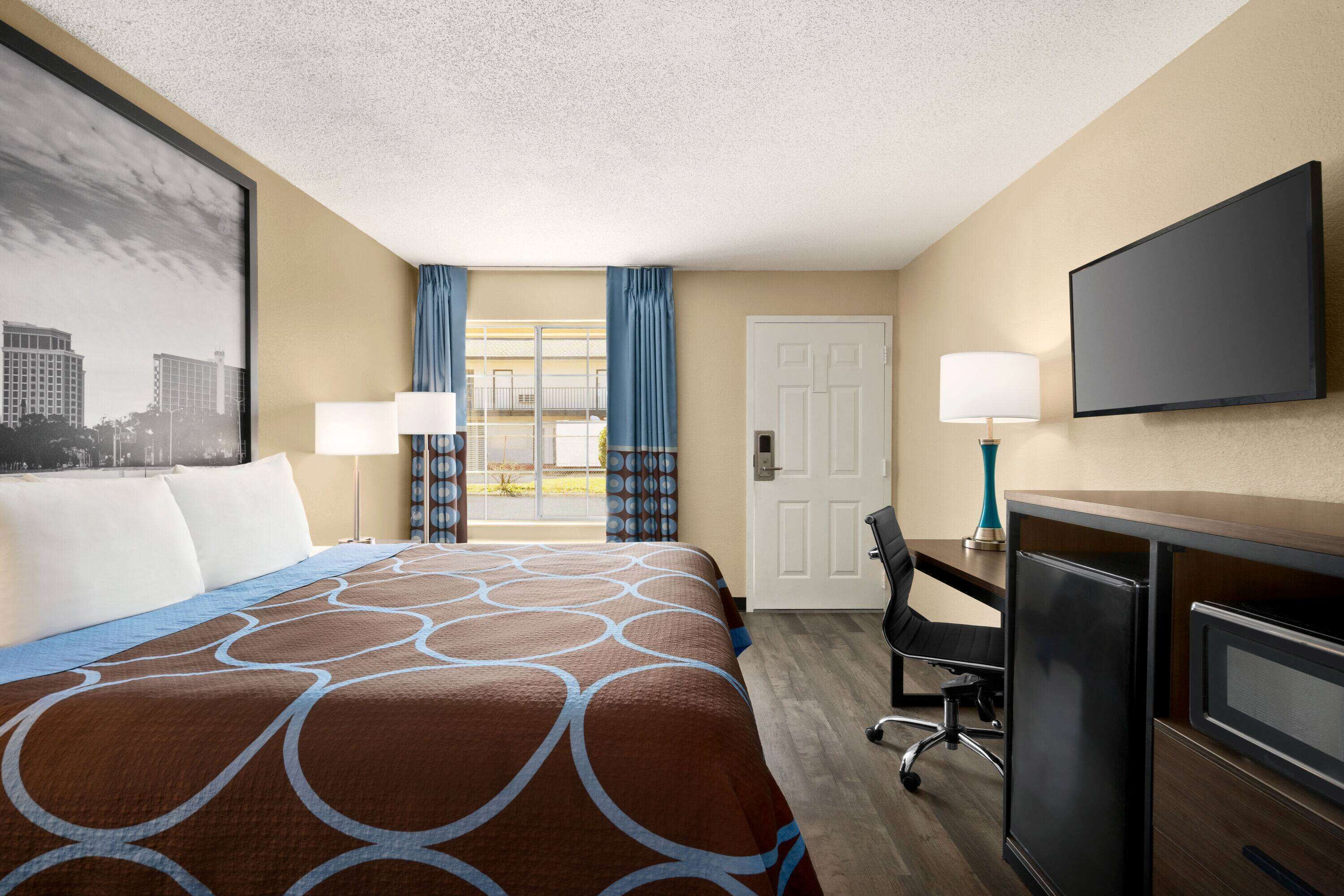 Habitación de hotel con una cama grande, un escritorio con computadora, televisión y ventana con cortinas.