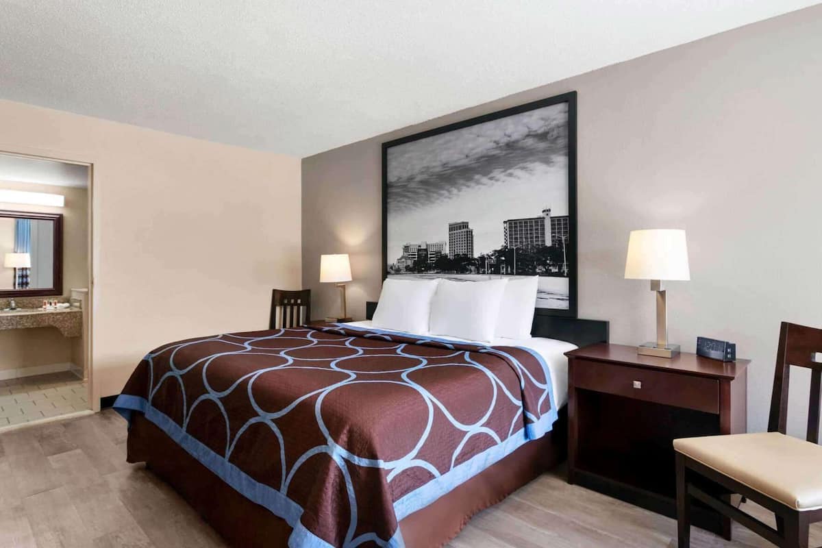 Habitación de hotel con cama, dos mesitas de noche, una silla, un cuadro grande con un paisaje urbano y un baño visible a través de una puerta abierta.