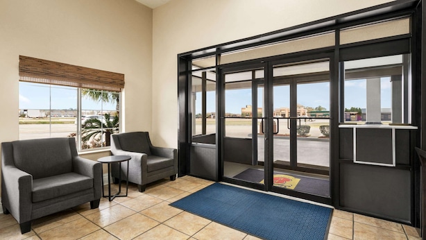A hotel lobby with a reception desk, two armchairs, a small table, and a large glass door leading outside. - Super 8 by Wyndham Gulfport Airport NW/Near Biloxi (Gulfport)
