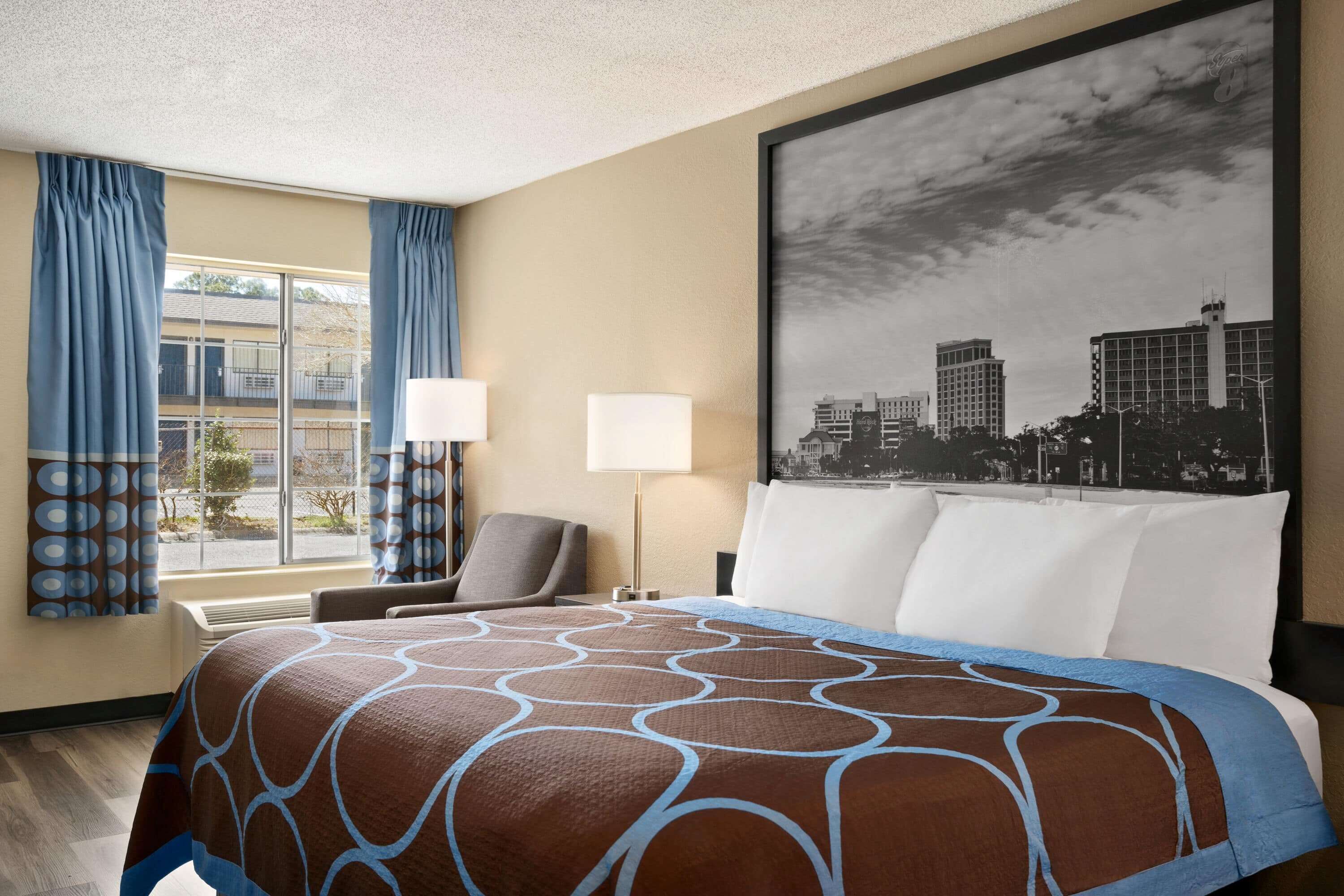 Ein Hotelzimmer mit einem großen Bett, einem Sessel, einem Schreibtisch und einem Fenster mit Vorhängen.