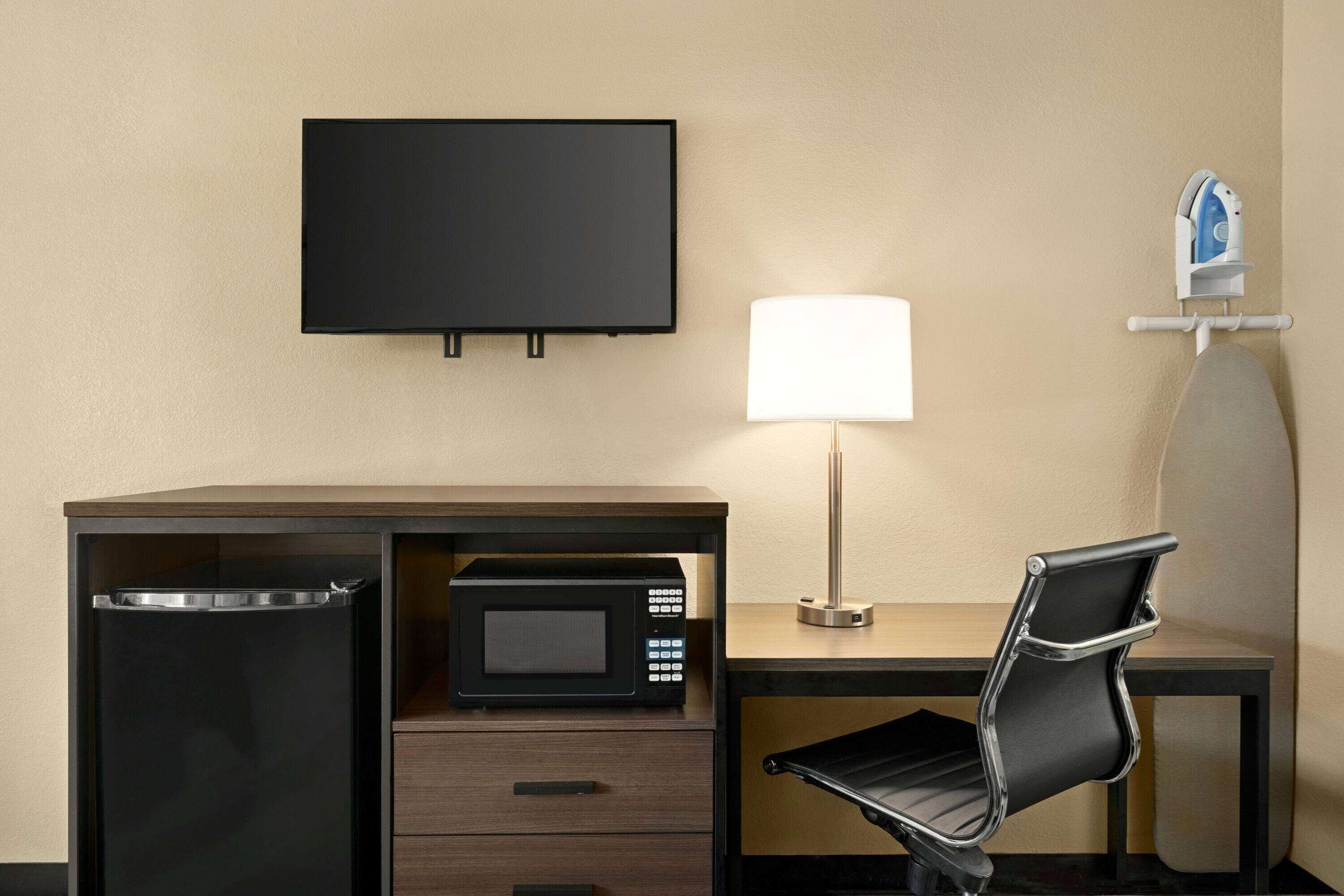 Habitación de hotel con televisión de pantalla plana, microondas, lámpara, silla y una tabla de planchar pequeña.