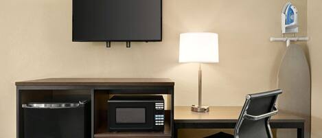 Habitación de hotel con televisión de pantalla plana, microondas, lámpara, silla y una tabla de planchar pequeña.