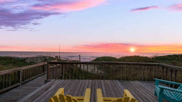 A wooden deck with two yellow Adirondack chairs facing a beach at sunset.