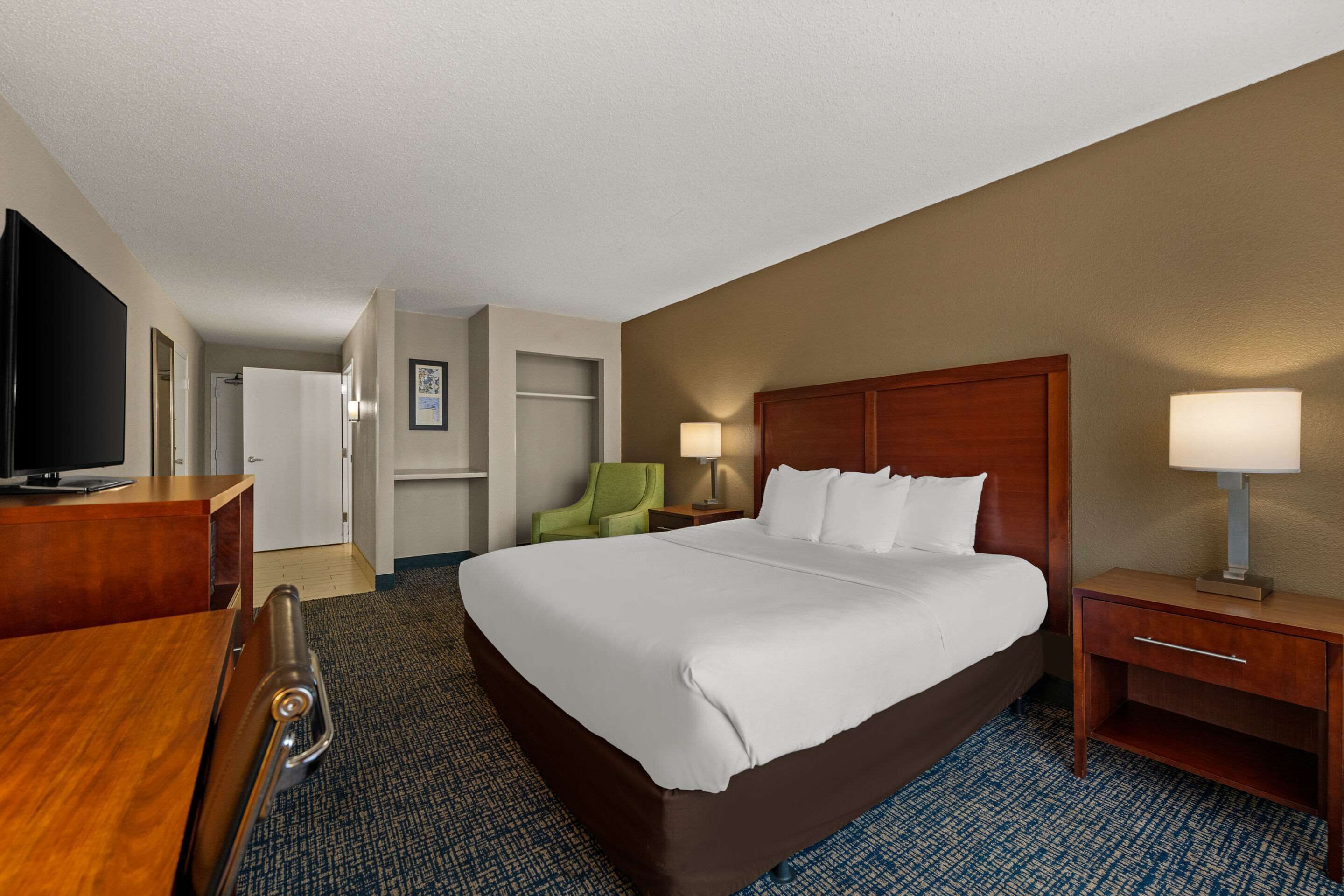 A hotel room with a large bed, a desk with a television, a chair, a nightstand with a lamp, and a wardrobe.