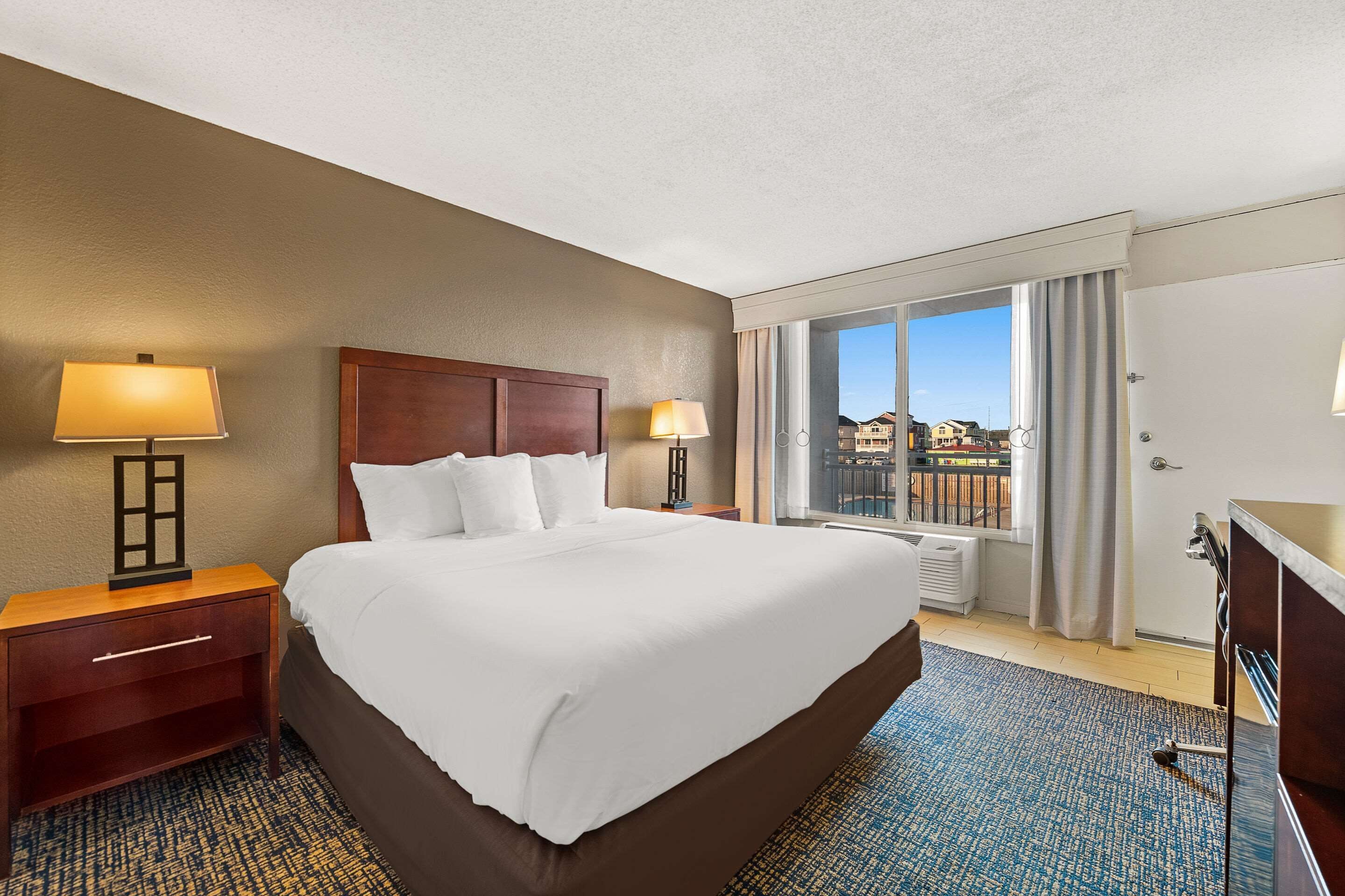 A hotel room with a large bed, two bedside tables with lamps, a dresser, and a view of buildings through a large window.