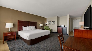 Une chambre d’hôtel comprenant un grand lit, un bureau, une chaise, une télévision, une lampe de bureau, une table de chevet, un fauteuil vert et une kitchenette.