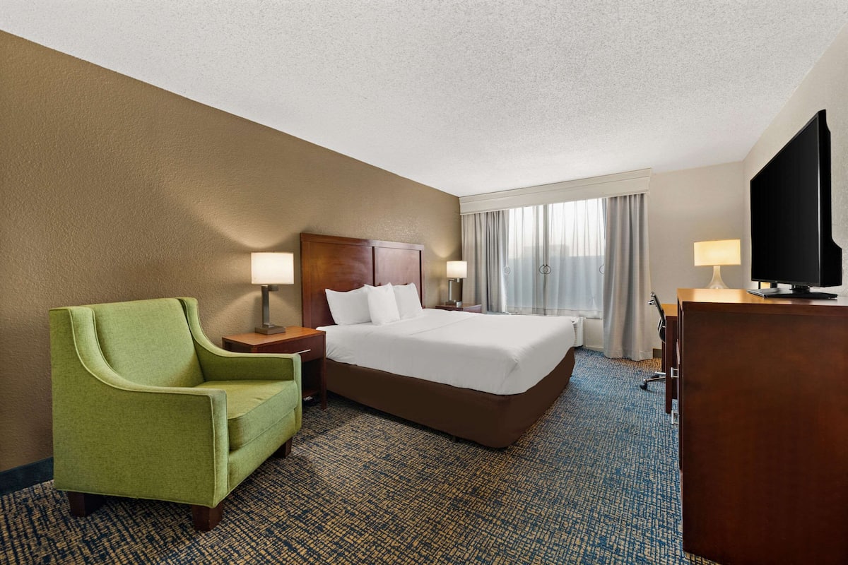 A hotel room with a bed, a green armchair, a desk with a lamp, a television, and a large window with curtains.