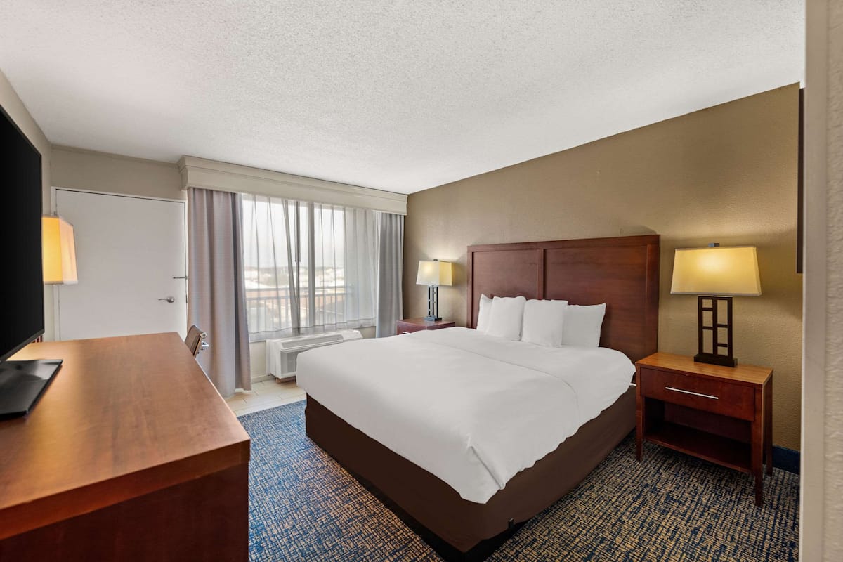 A hotel room with a large bed, a desk, a chair, a nightstand with a lamp, and a window with curtains.