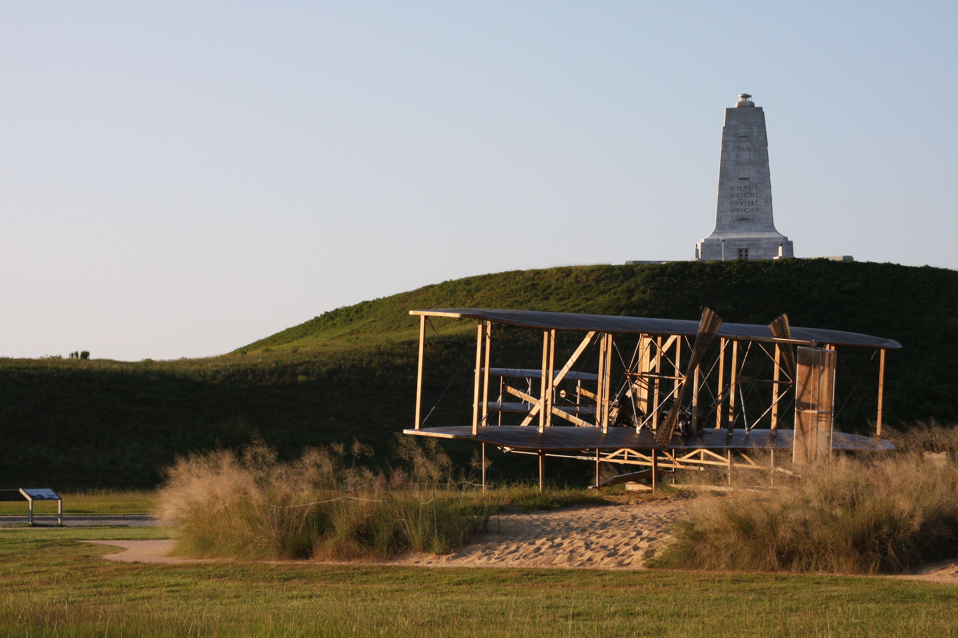 Une structure en bois surmontée d’un toit métallique, située sur une colline herbeuse, avec un monument en arrière-plan et un ciel dégagé.