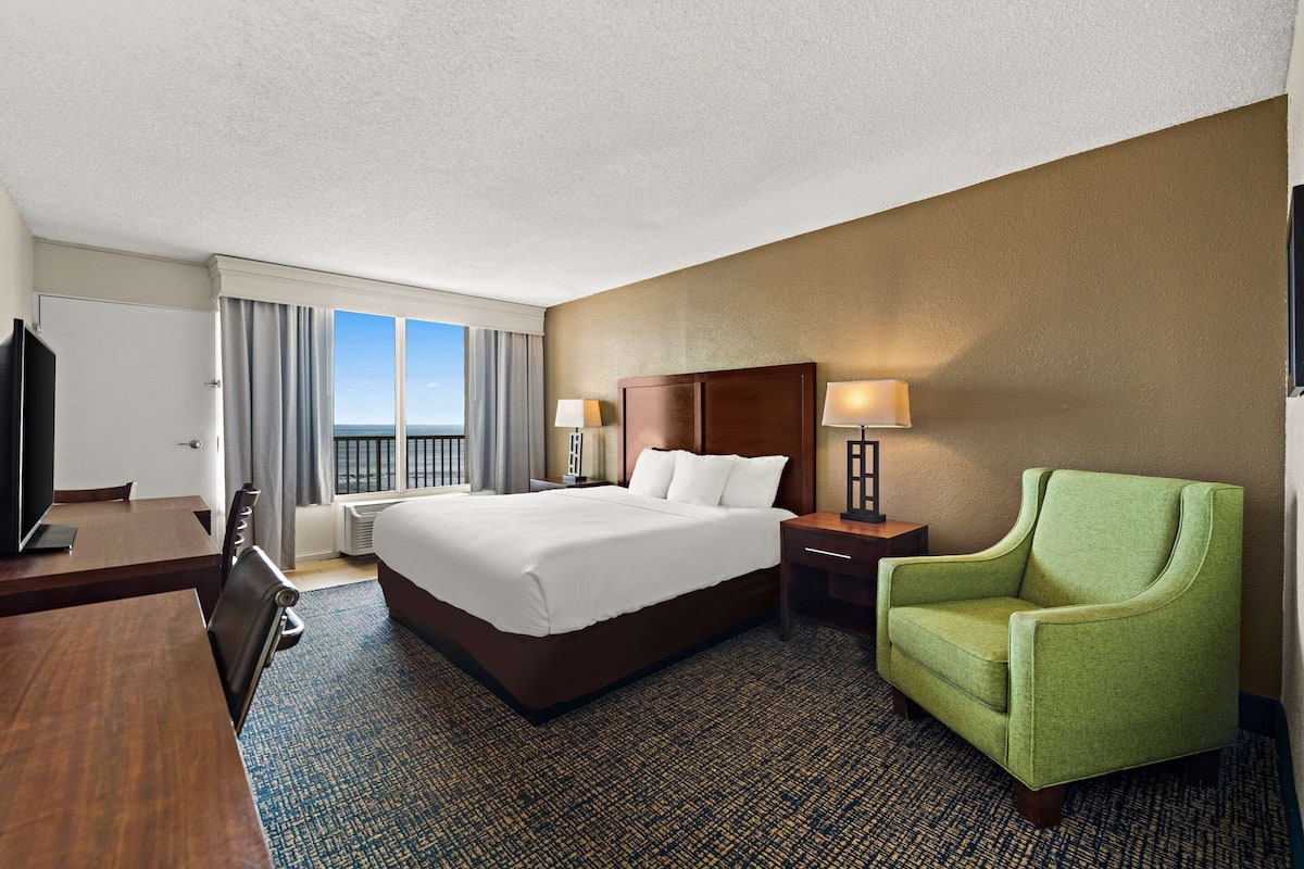 A hotel room with a large bed, a green armchair, a desk with a computer, and a view of the ocean.