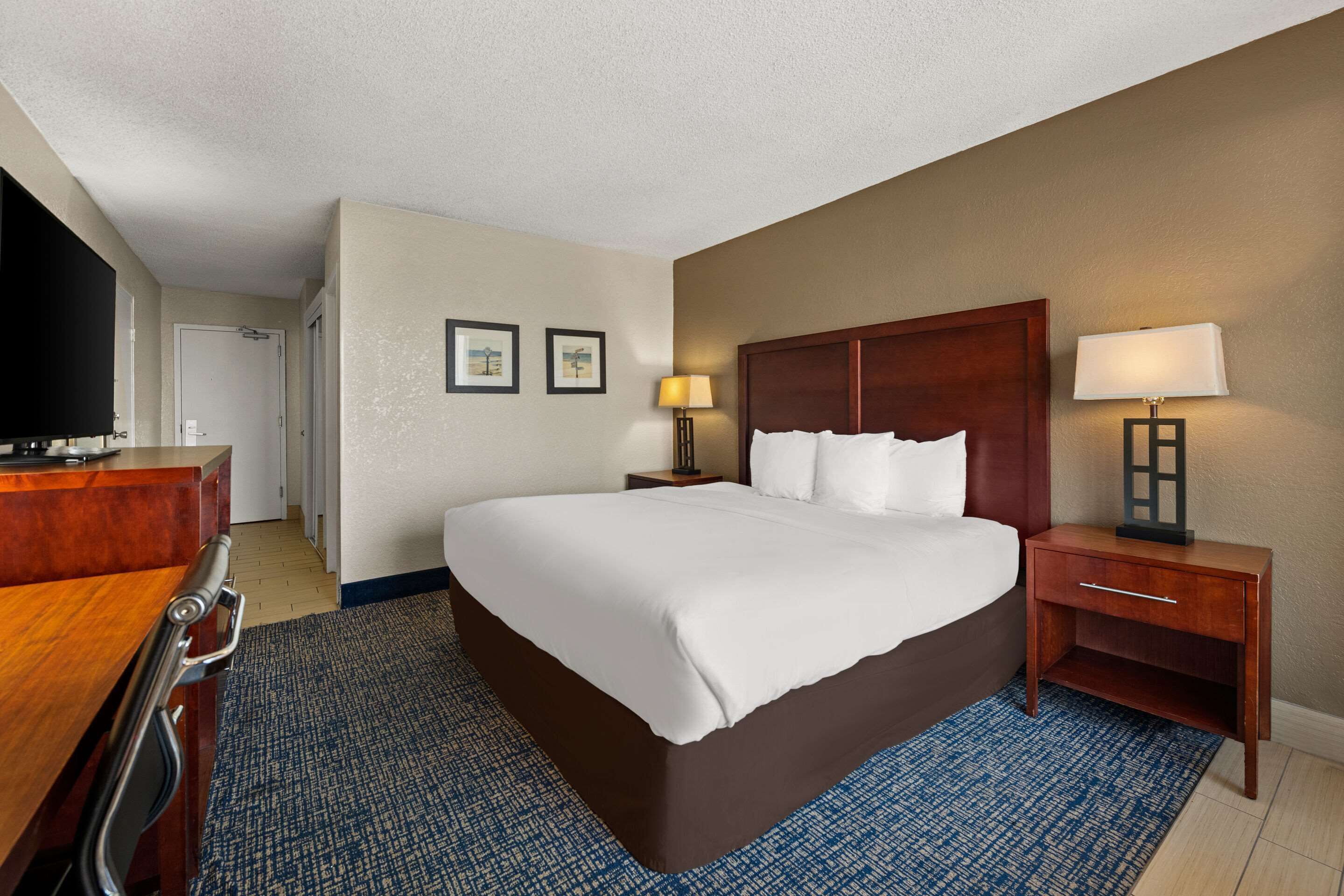 A hotel room with a large bed, a desk with a chair, a television, two bedside lamps, and a blue patterned rug.