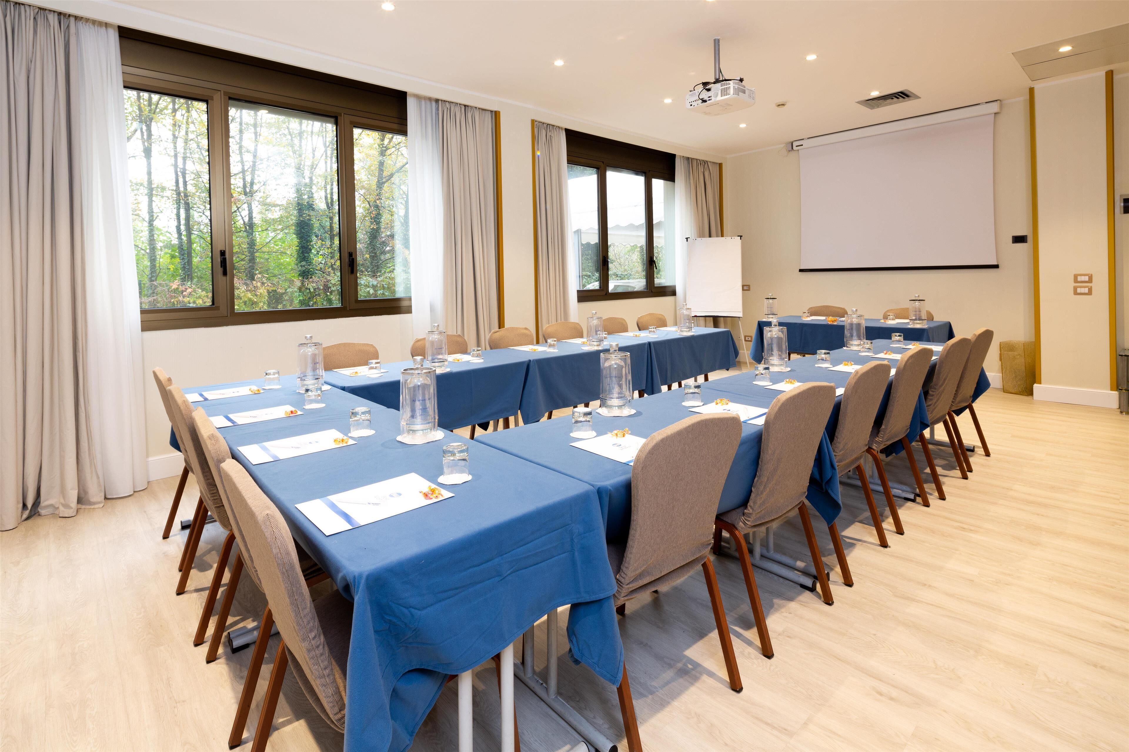 A conference room with a long table, chairs, a projector screen, and windows with curtains.