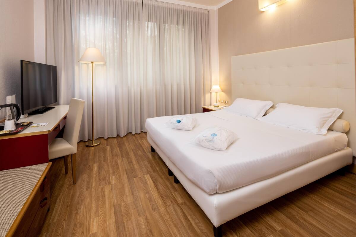 A hotel room with a bed, a desk with a TV, a chair, a lamp, and a bedside table.