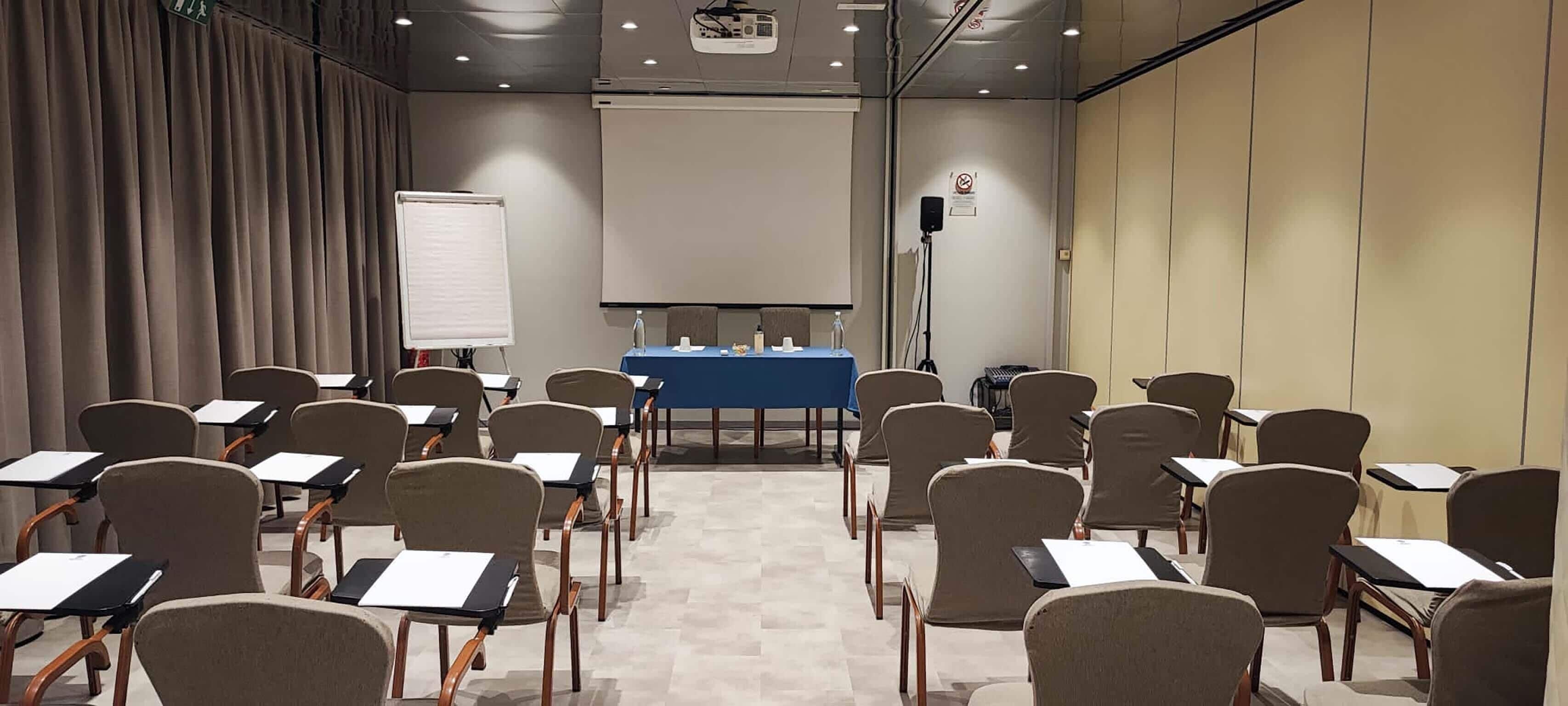 Un salón de conferencias con filas de sillas, una pantalla de proyector y una pizarra.