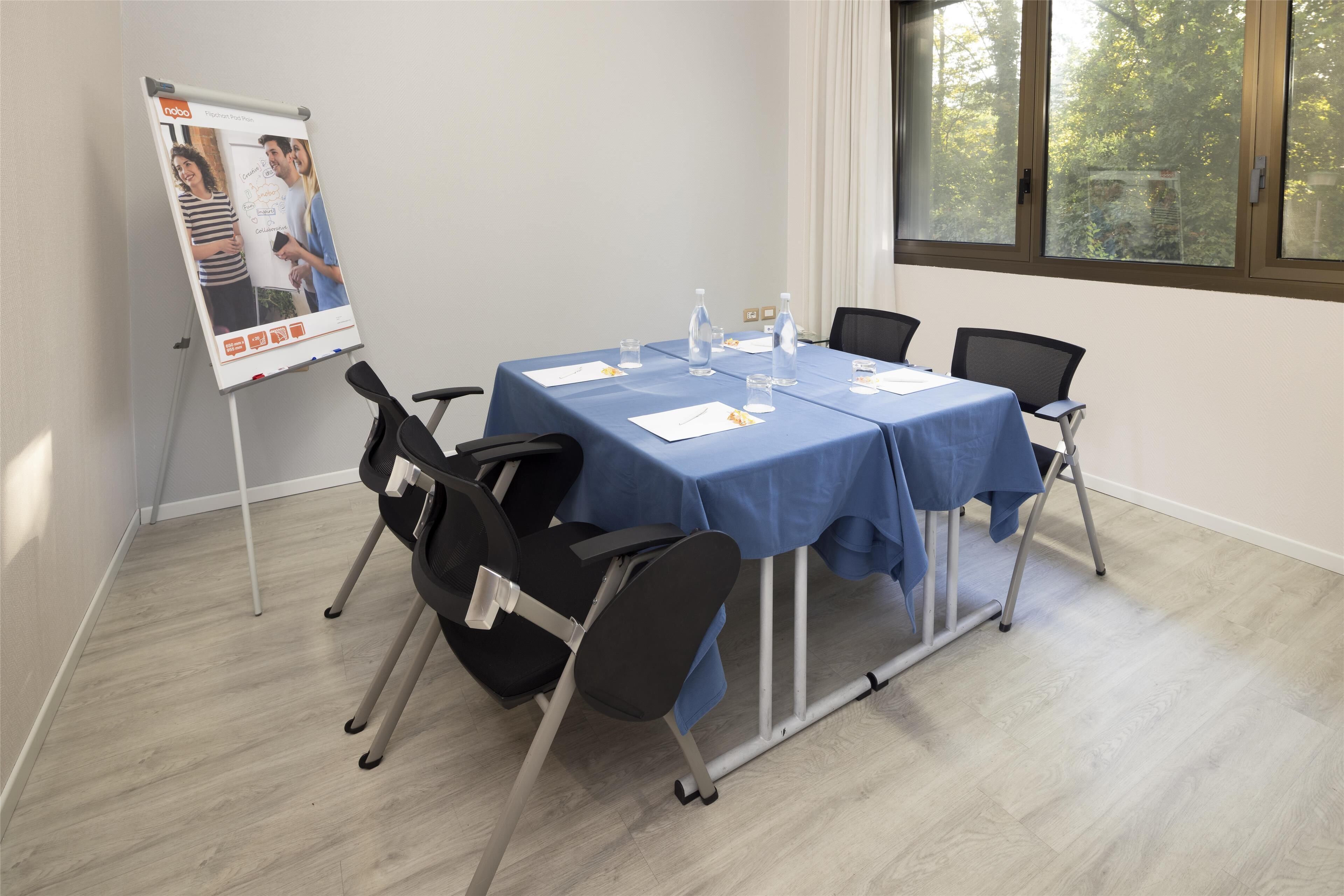 A conference room with a blue tablecloth, four chairs, a flip chart, and a window with a view of trees.