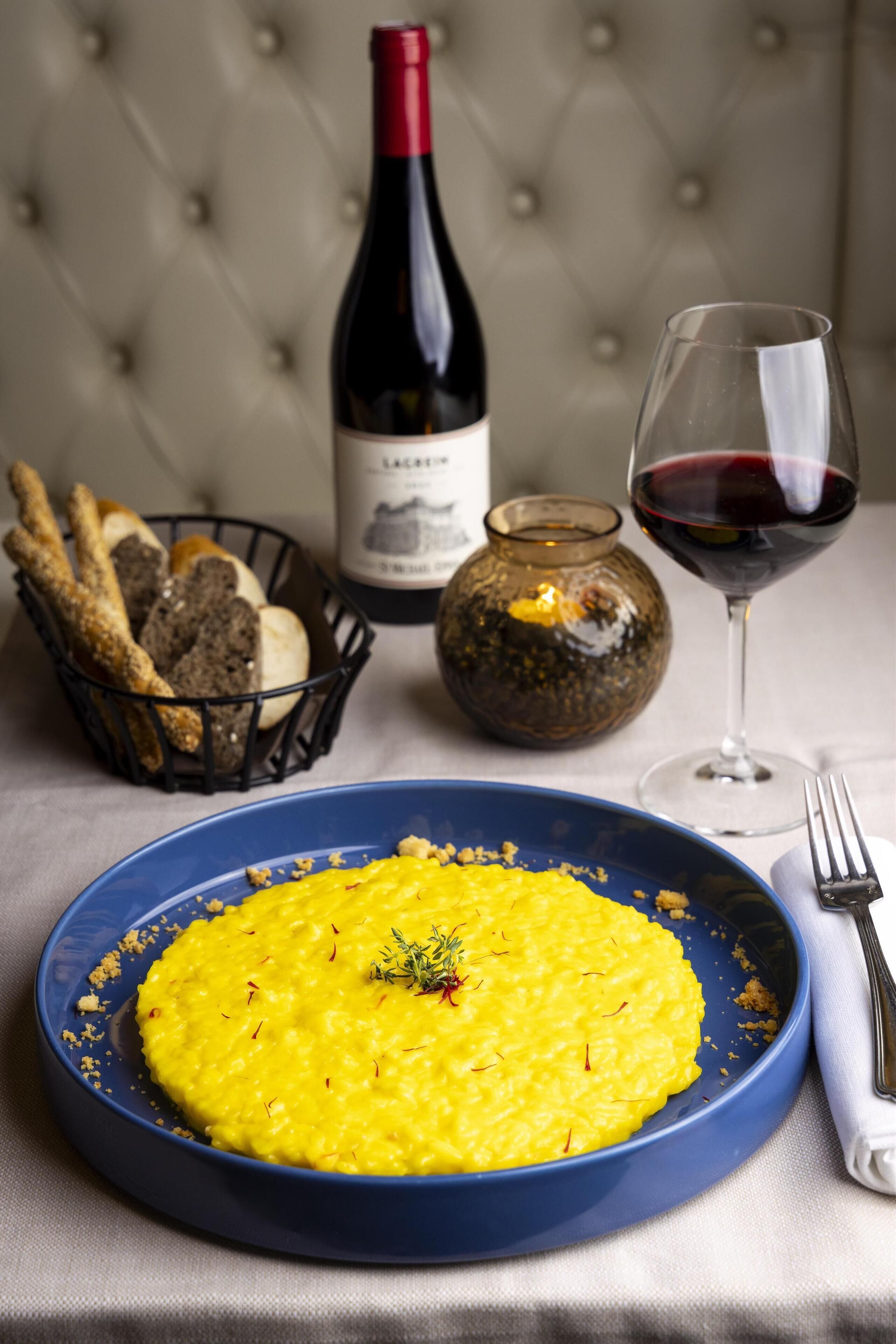 Un plato de risotto amarillo adornado con hierbas, una botella de vino, una copa de vino tinto y un cesto de palitos de pan.