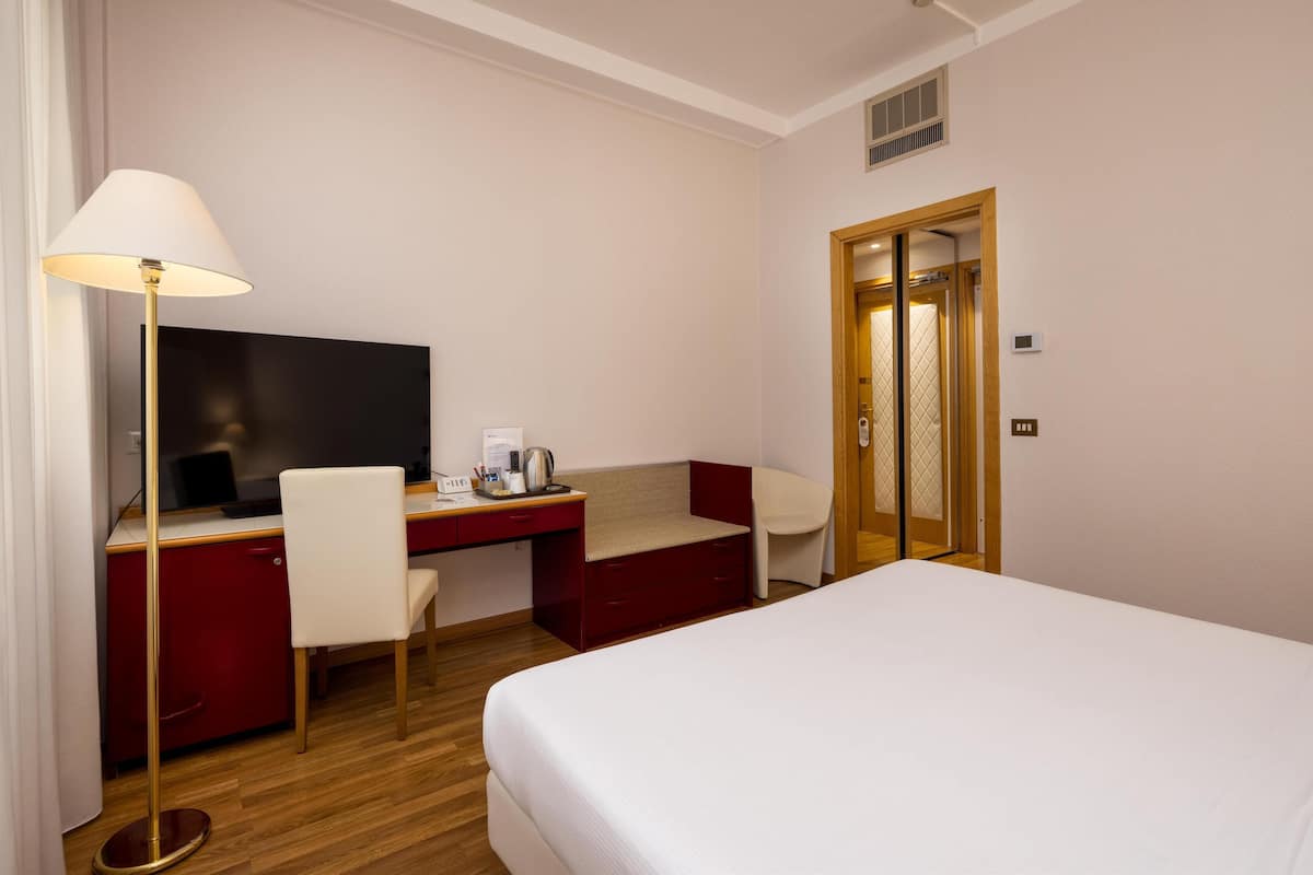 A hotel room with a bed, a desk with a computer, a chair, a lamp, and a television.