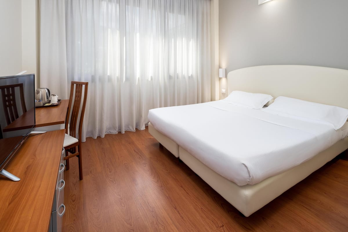 A hotel room with a bed, a desk with a kettle, a chair, and a window with curtains.