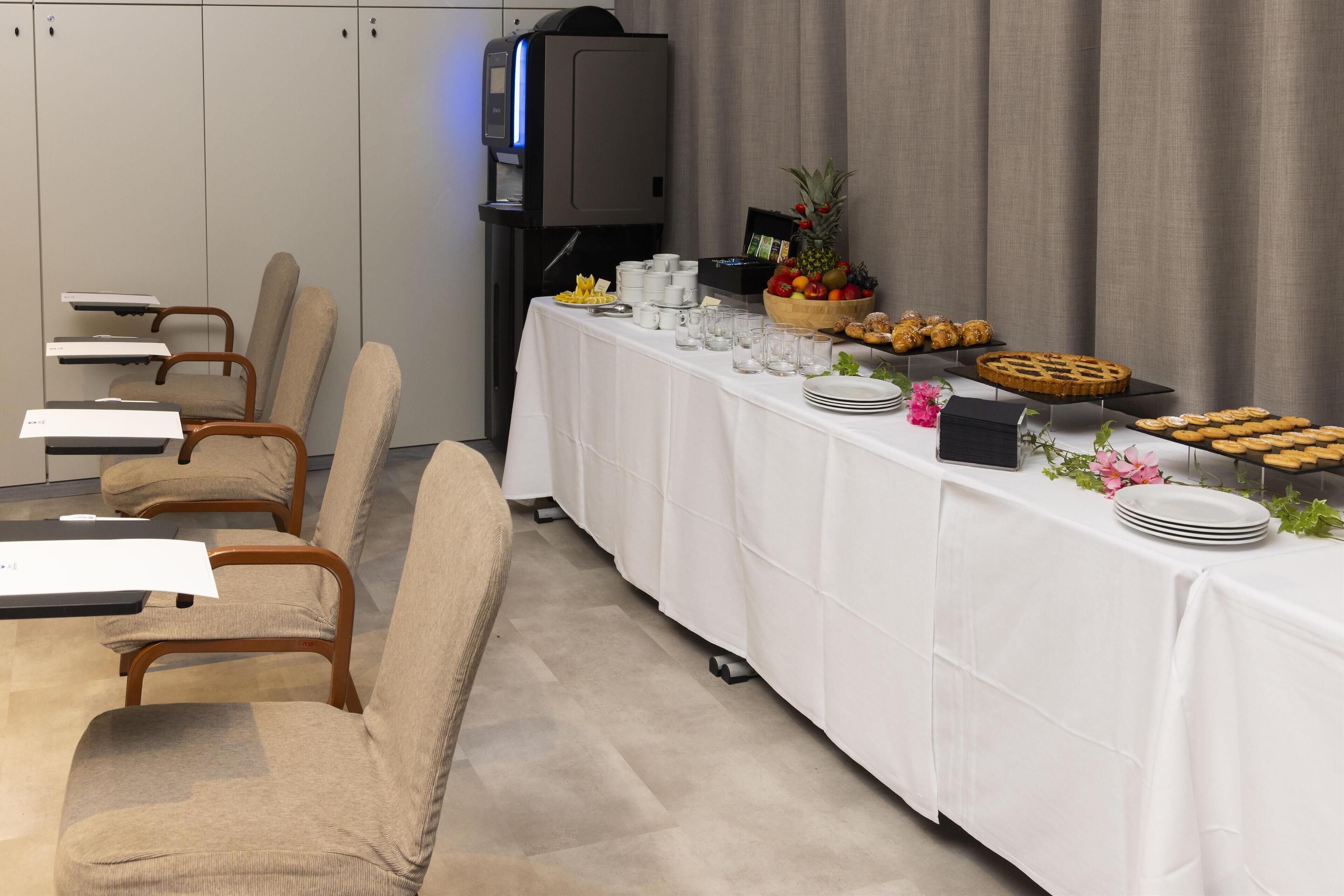 Un salón de conferencias con una larga mesa dispuesta para una reunión, equipado con sillas, un proyector y una estación de café.
