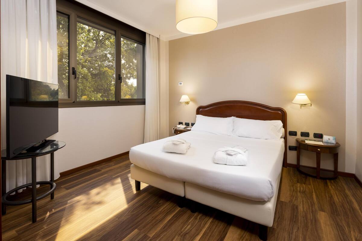 A hotel room with a large bed, two bedside lamps, a TV on a stand, and a window with curtains.
