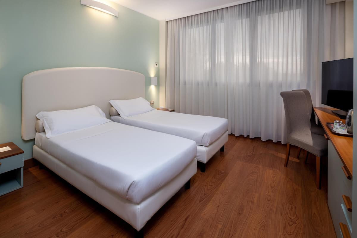 A hotel room with two single beds, a desk with a chair, a TV, and a large window with curtains.