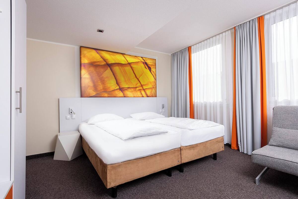 Une chambre d’hôtel avec un lit double, un fauteuil gris, un grand tableau abstrait et une fenêtre avec des rideaux orange.