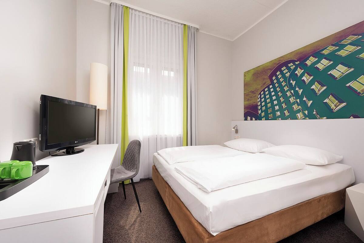 Une chambre d’hôtel avec un lit, un bureau équipé d’un ordinateur, une chaise et un tableau accroché au mur.