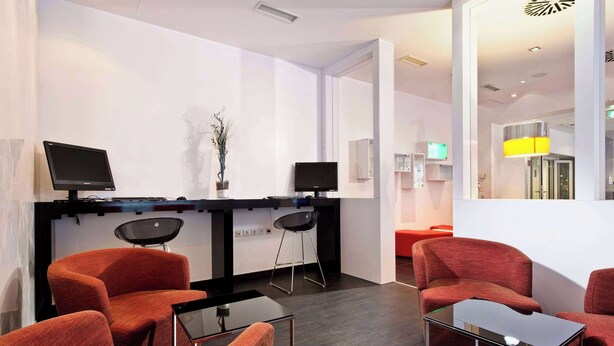 A modern interior with a reception desk, two monitors, a potted plant, and red upholstered chairs. - Wyndham Garden Düsseldorf City Centre Königsallee (Düsseldorf)