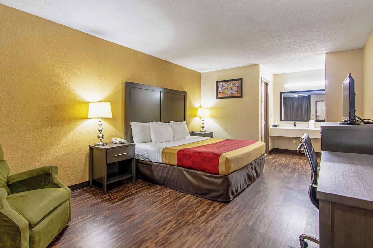 A hotel room with a bed, two bedside tables with lamps, a green armchair, a desk with a computer, and a large window.