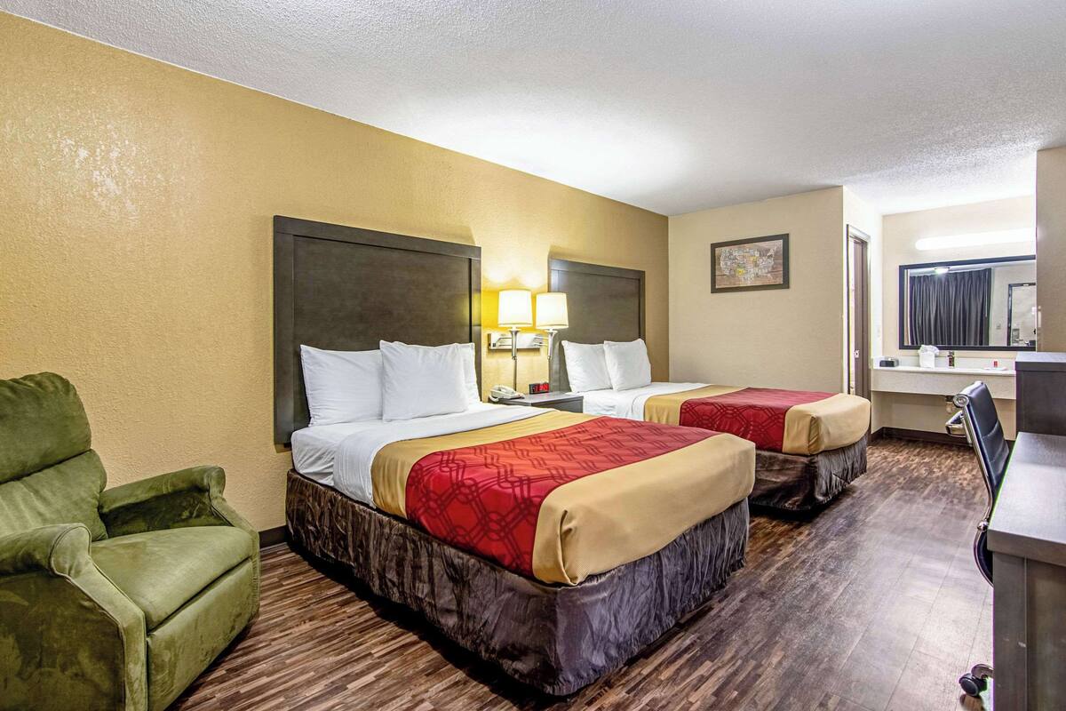 A hotel room with two beds, a green armchair, a desk, and a mirror.