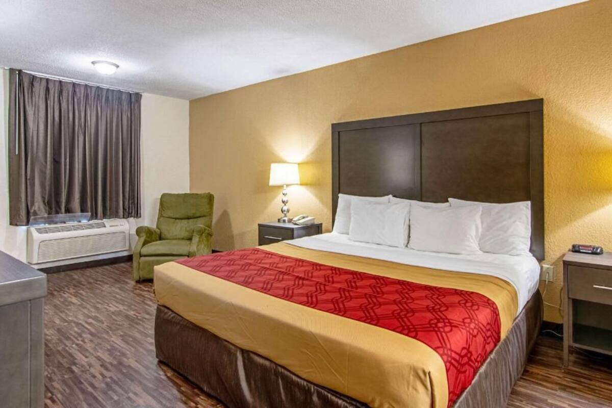 A hotel room with a bed, a chair, a nightstand, a lamp, and an air conditioning unit.