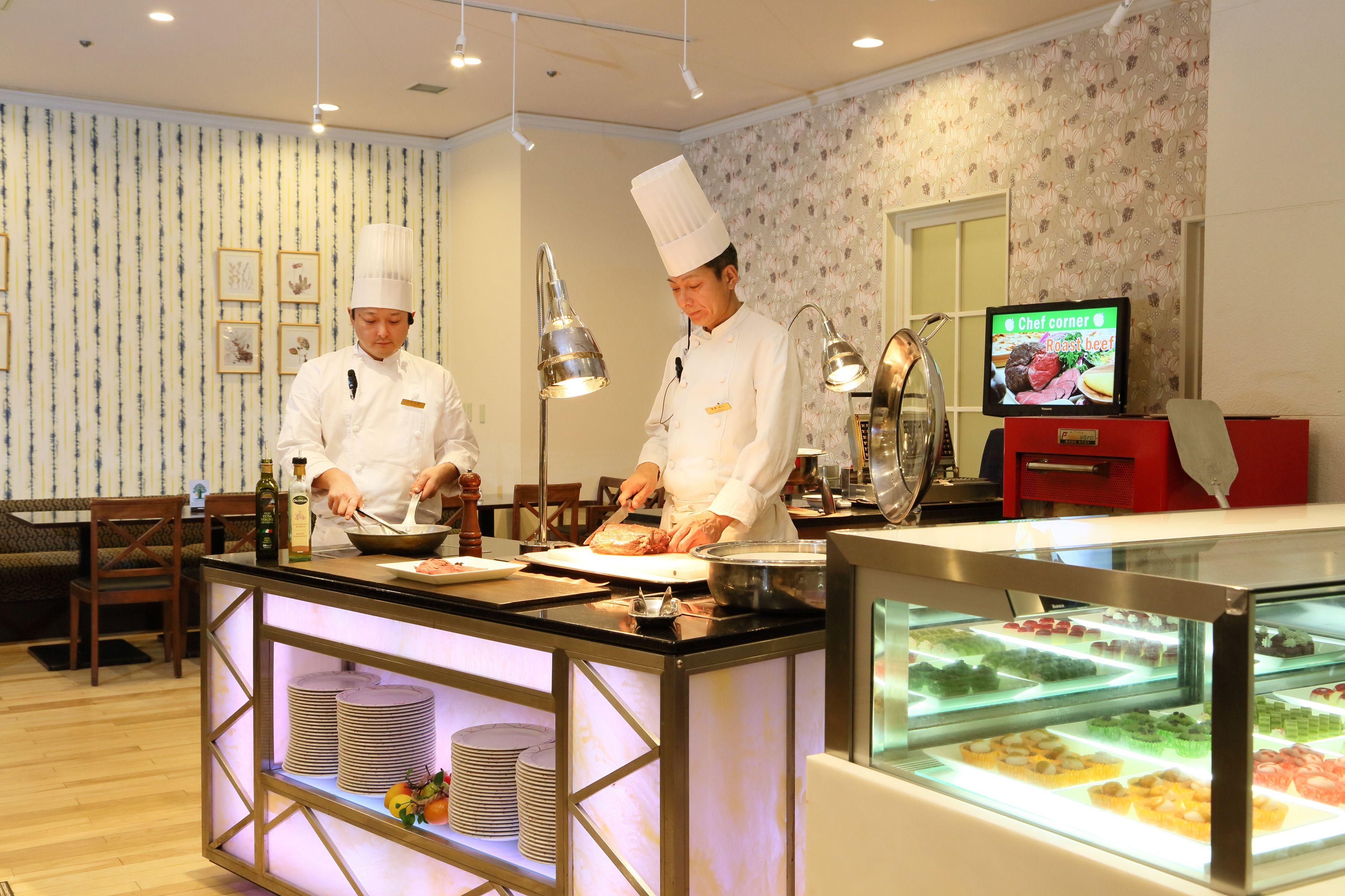 A kitchen area with chefs preparing food, a display case with desserts, and a television displaying a cooking program.