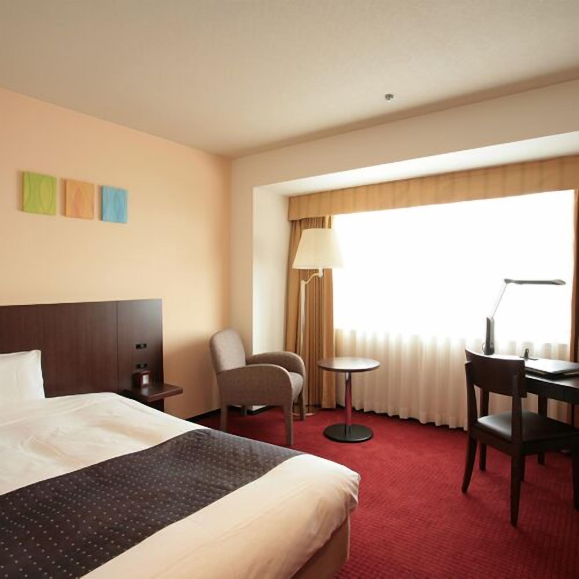 Habitación de hotel con una cama, un escritorio con silla, una lámpara y una ventana con cortinas.