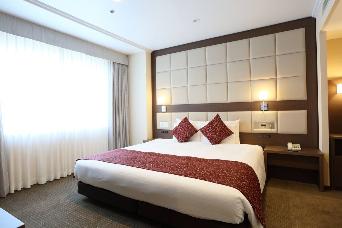 Habitación de hotel con una cama grande, cabecera con panel de luz, mesita de noche con teléfono y ventana con cortinas.