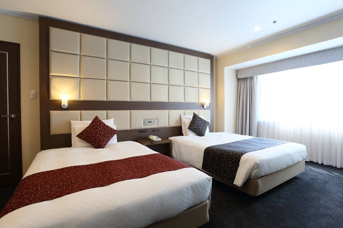 Habitación de hotel con dos camas, cabecera con panel de luz, mesita de noche con teléfono y ventana con cortinas.