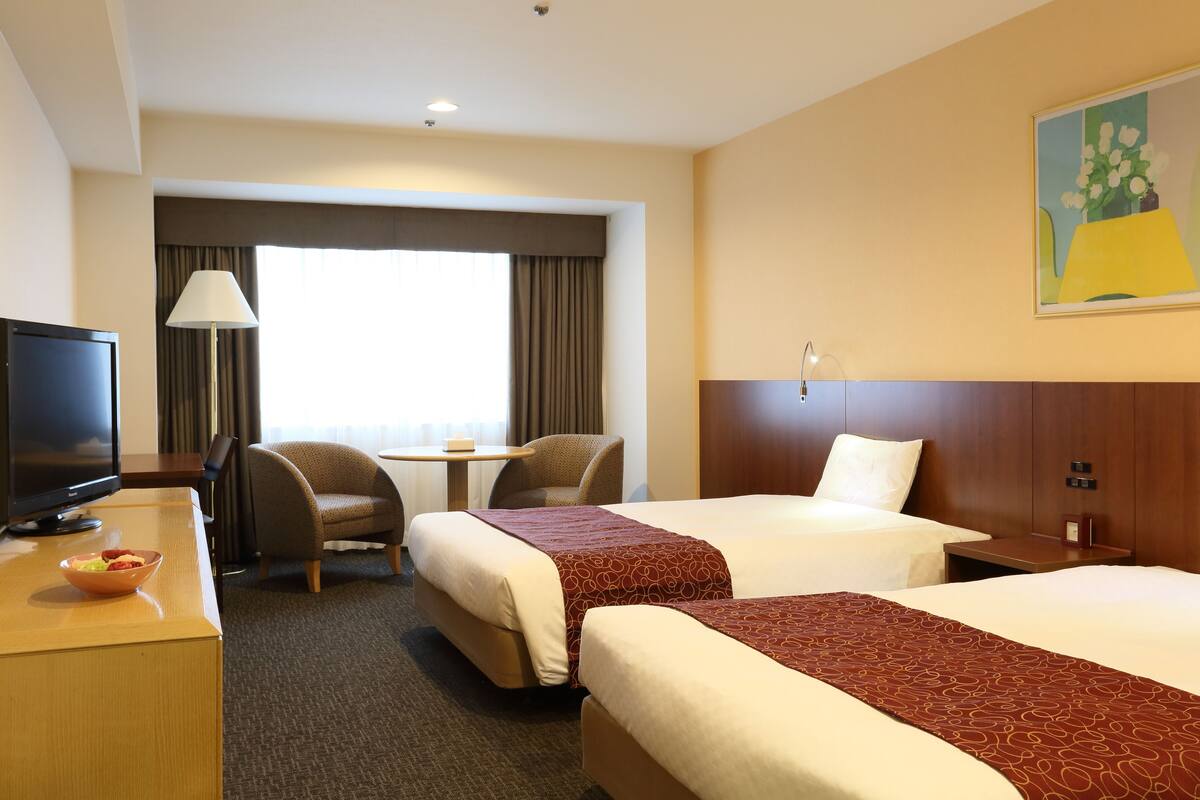 Habitación de hotel con dos camas, un escritorio con televisor, una silla, una lámpara y un cuadro en la pared.