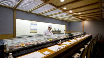 A sushi bar with a chef behind the counter, a glass display case with various food items, and a seating area with chairs.