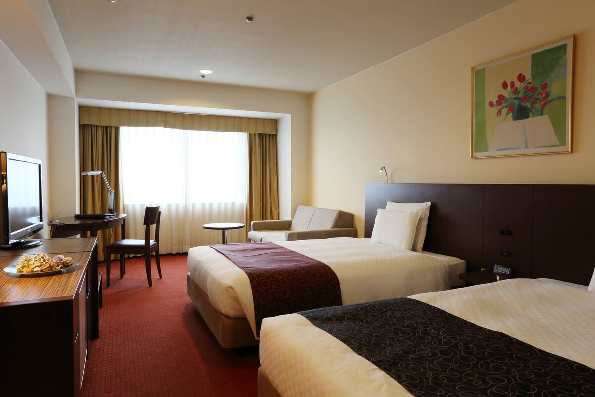 Habitación de hotel con dos camas, un escritorio con silla, televisión y ventana con cortinas.