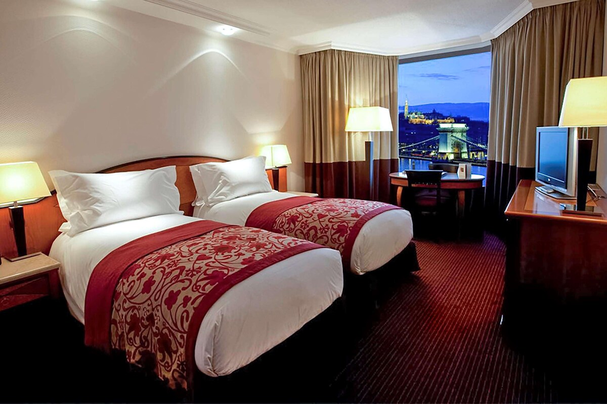 A hotel room with two beds, a desk, a television, and a view of a cityscape at dusk.