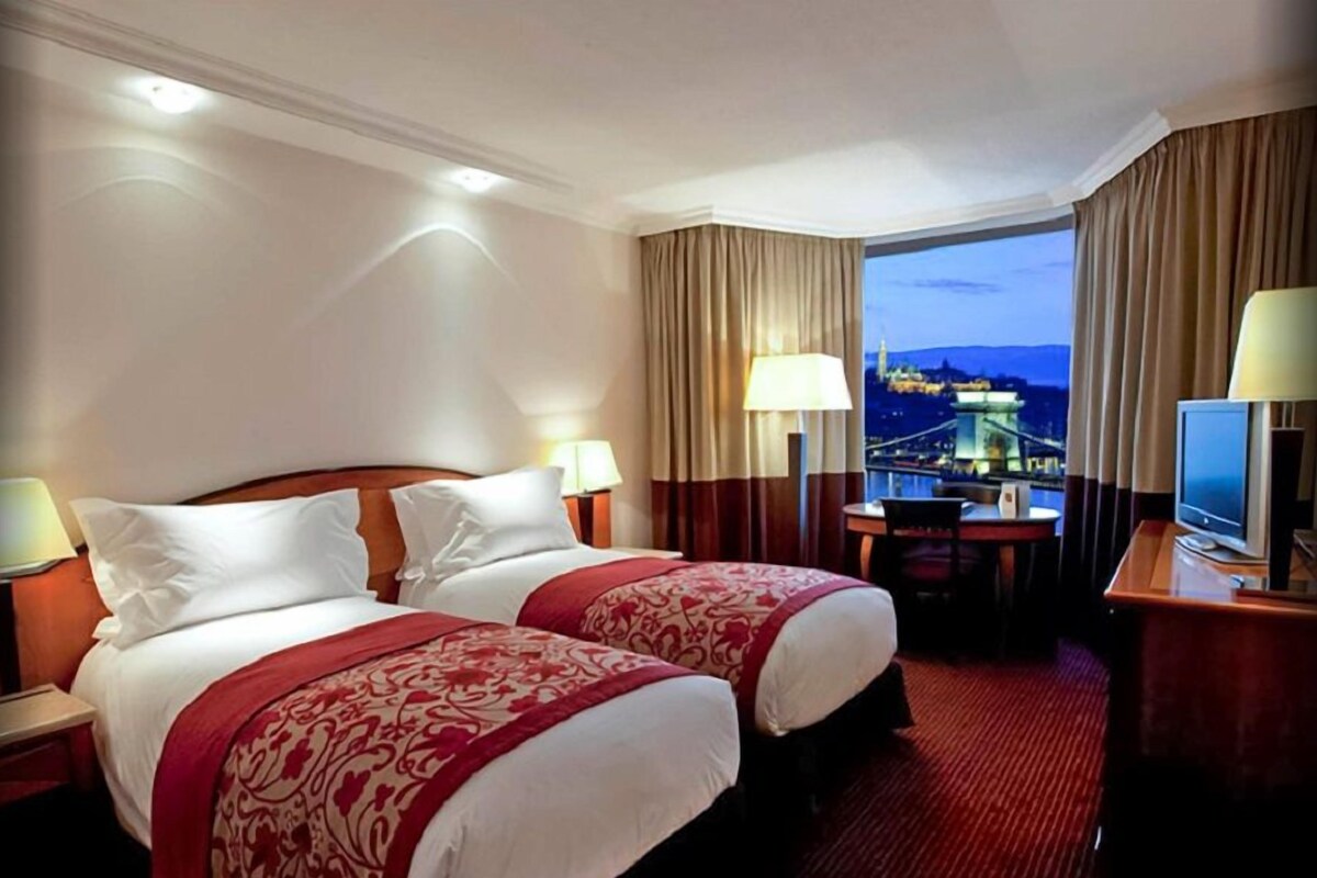 A hotel room with two beds, a desk, a chair, and a TV. There is a window with a view of a bridge and city lights.
