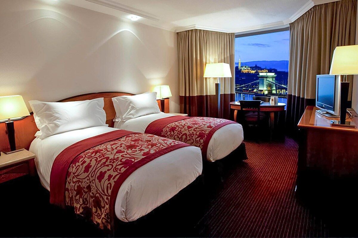 A hotel room with two beds, a desk, a television, and a view of a cityscape at dusk.
