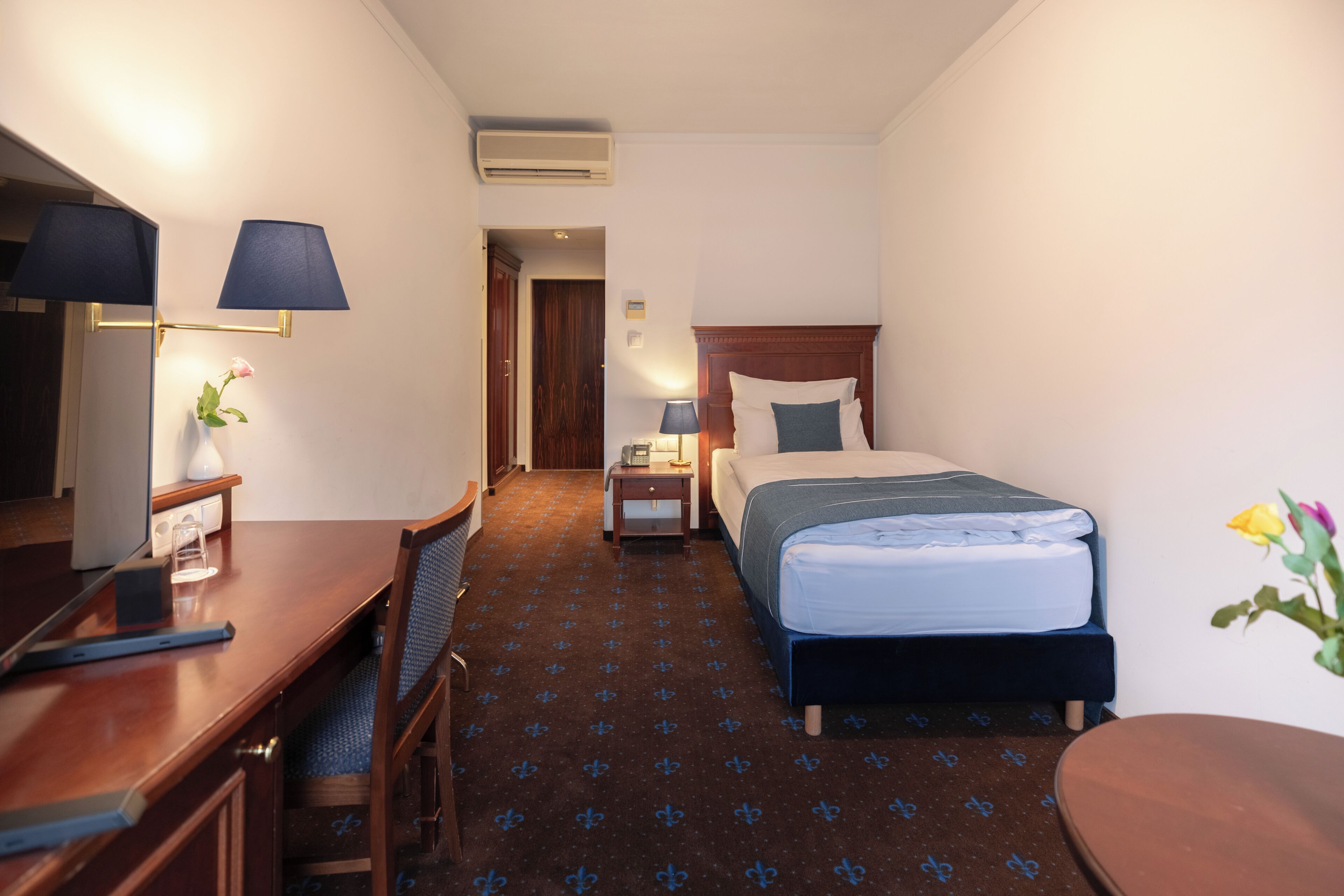 Camera d'albergo con un letto, una scrivania, una sedia, un comodino, una lampada e un tavolino con un vaso di fiori.