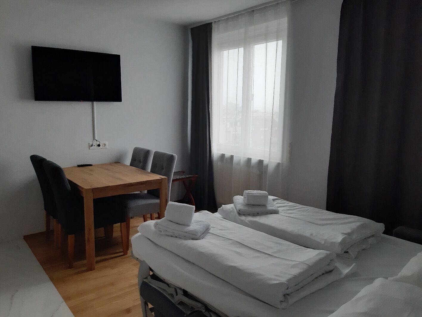 A hotel room with a wooden dining table, two beds with white linens, a flat-screen TV mounted on the wall, and a window with sheer curtains.