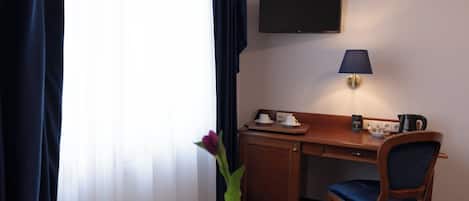 Habitación de hotel con escritorio, silla, televisor, lámpara y un florero con una flor.