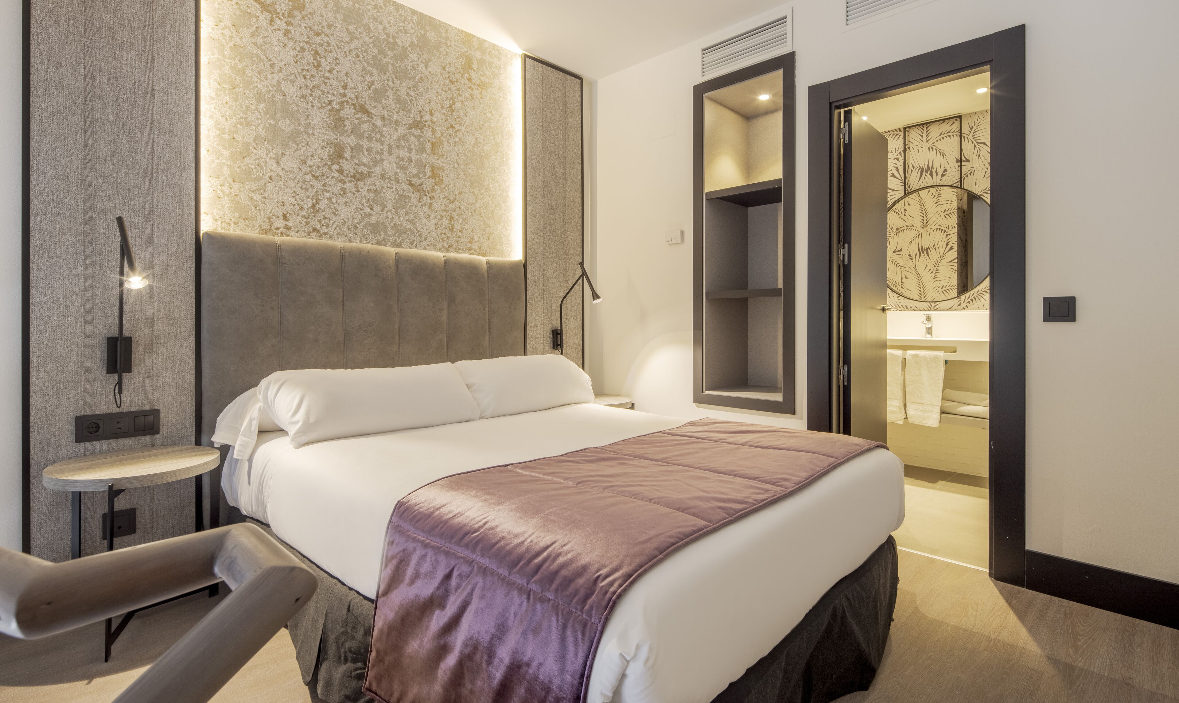A modern hotel room with a large bed, a small bedside table, a chair, and a bathroom visible through an open door.